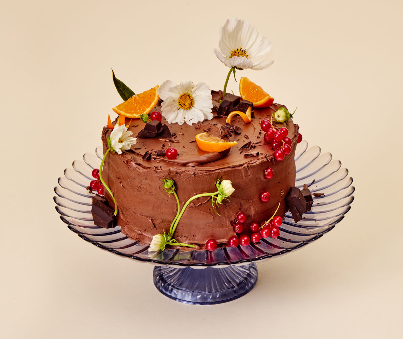 Chocolate cake with fruit and flowers on a glass cake stand against a beige background