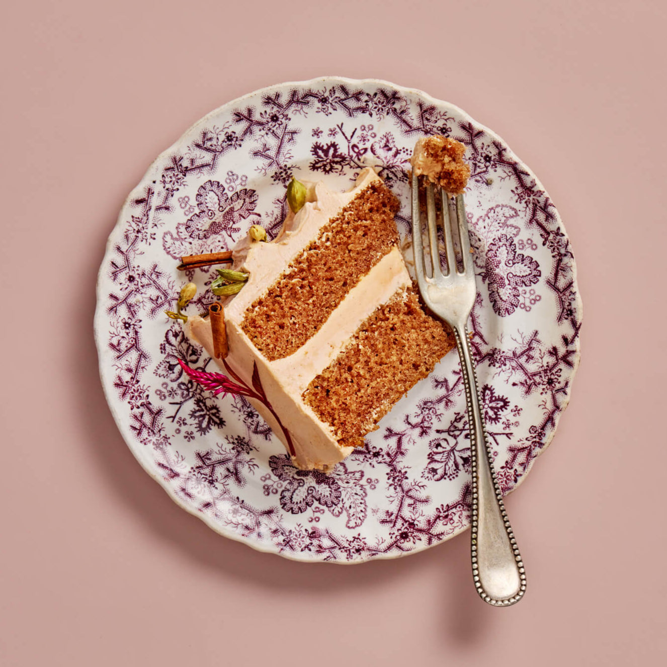 Slice of layered cake on a decorative plate with a fork, against a pink background