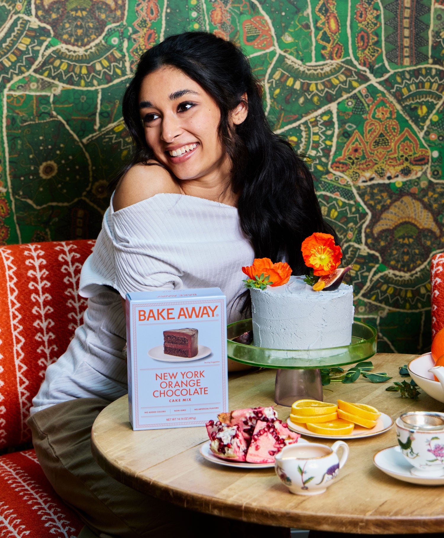 Woman sitting at a table with a cake and 'Bake Away' book against a colorful wall.