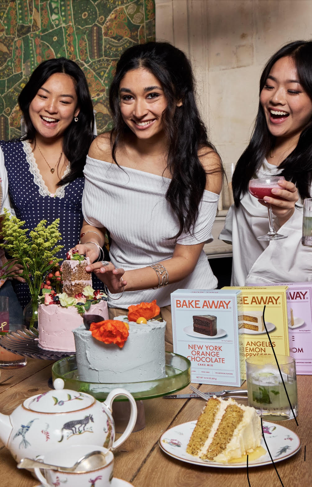 Group of women gathered around a table with cakes and drinks, smiling and enjoying each other's company.