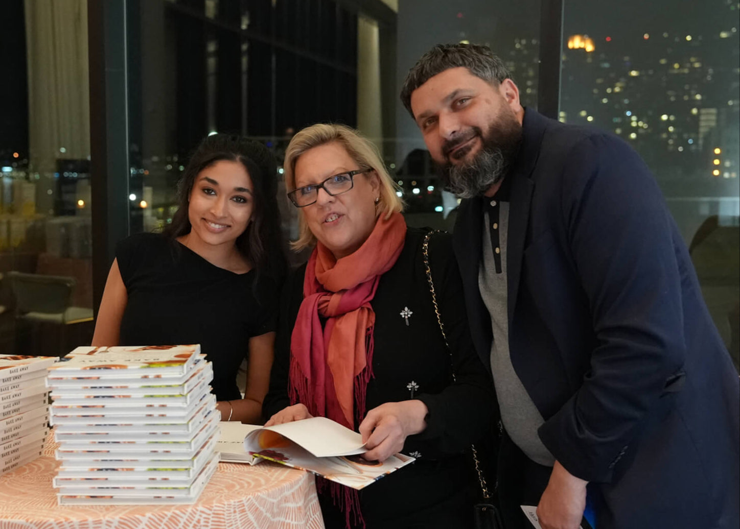 Three people standing around a table with books, with a cityscape in the background.