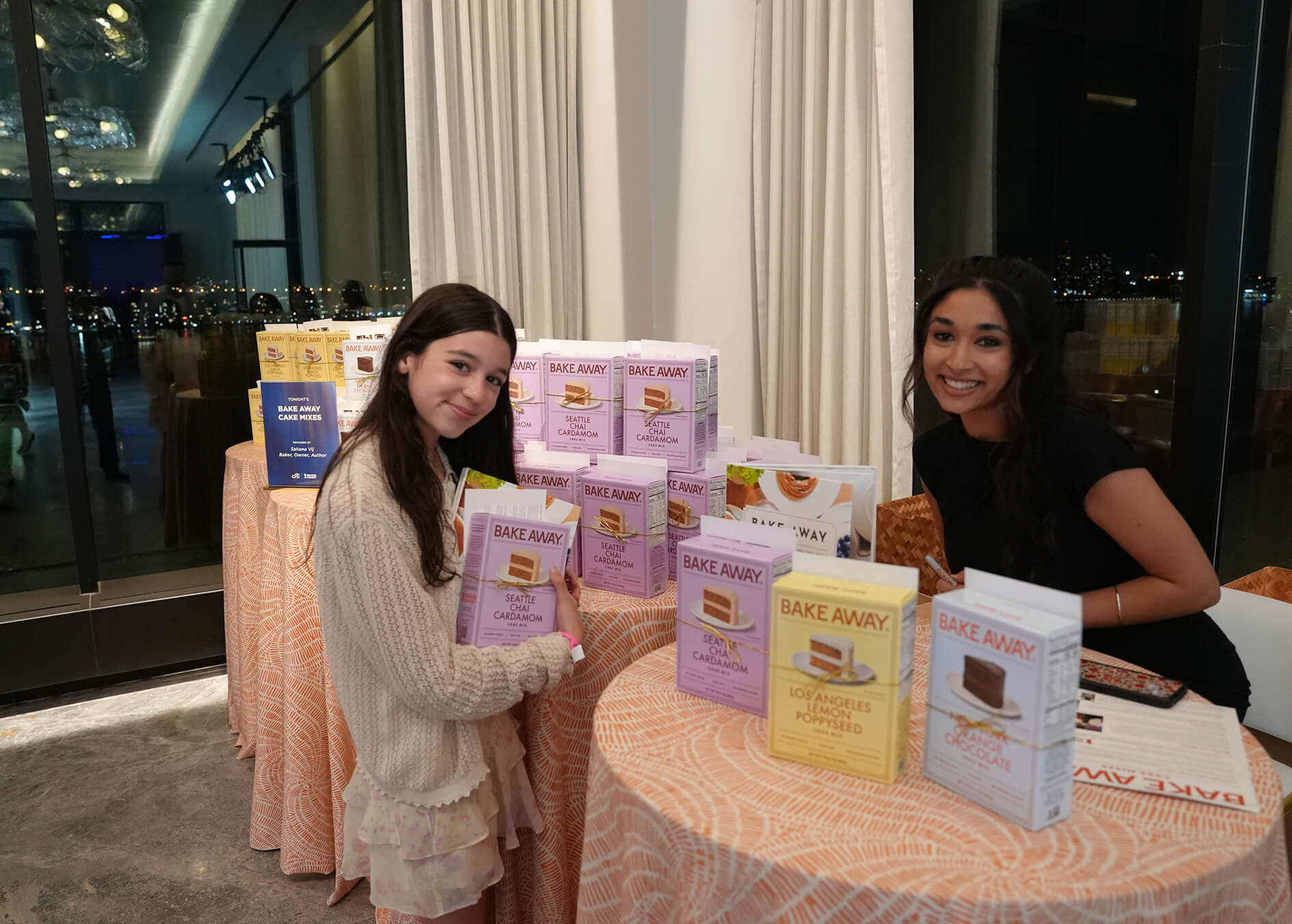 Two women standing behind a table displaying various boxes of 'Bake Away' products in an indoor setting.