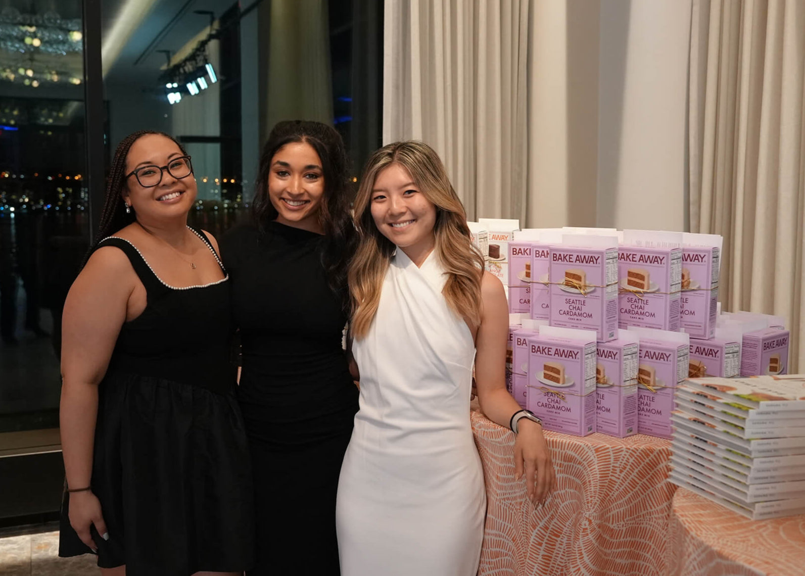 Three women posing together with a table displaying boxes and stacks of paper products.