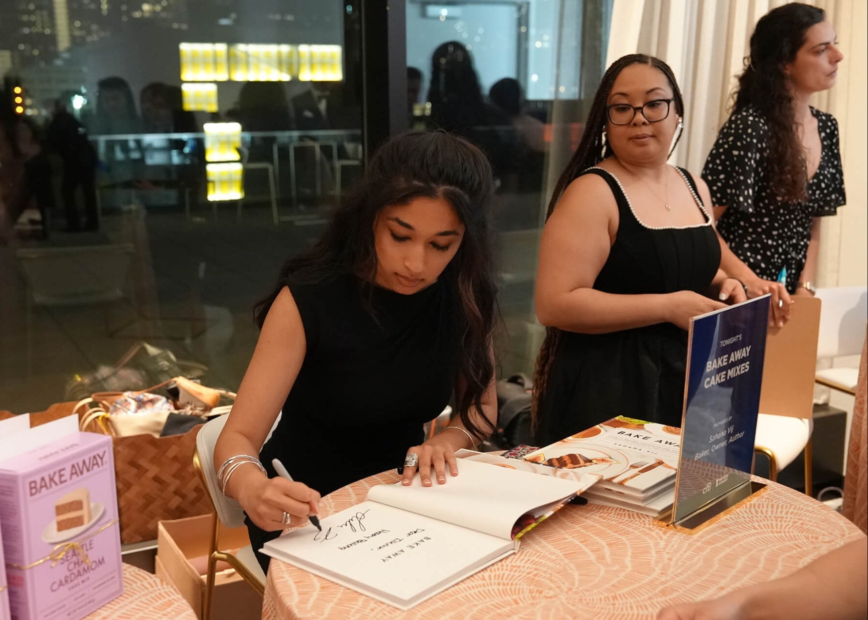 Two women at a table with a book and other items, possibly at an event or gathering.
