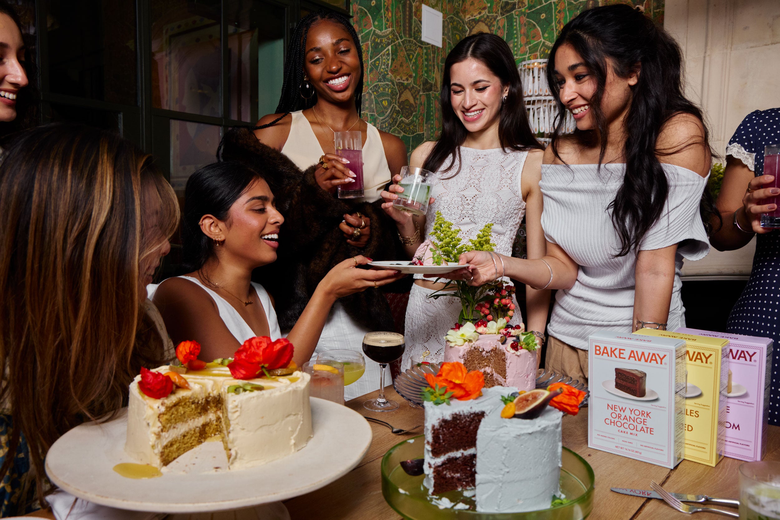 Group of women gathered around a table with cakes and drinks, celebrating.