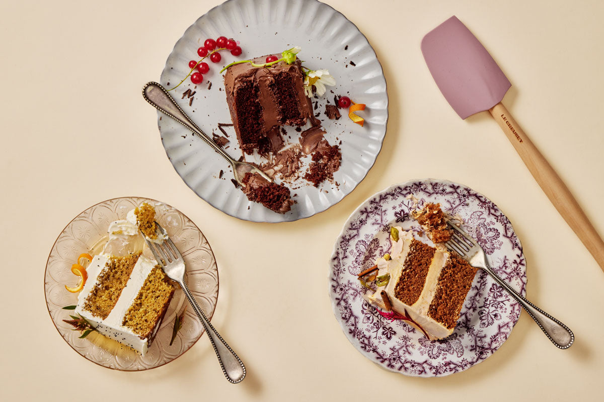 Three slices of cake on decorative plates with a Le Creuset spatula on a beige background