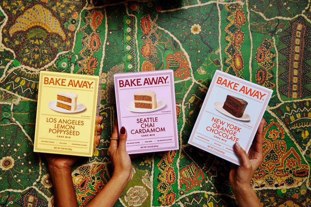 Three 'Bake Away' baking mix boxes held by hands on a patterned fabric background