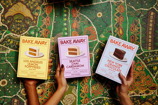Three 'Bake Away' baking mix boxes held by hands on a patterned fabric background