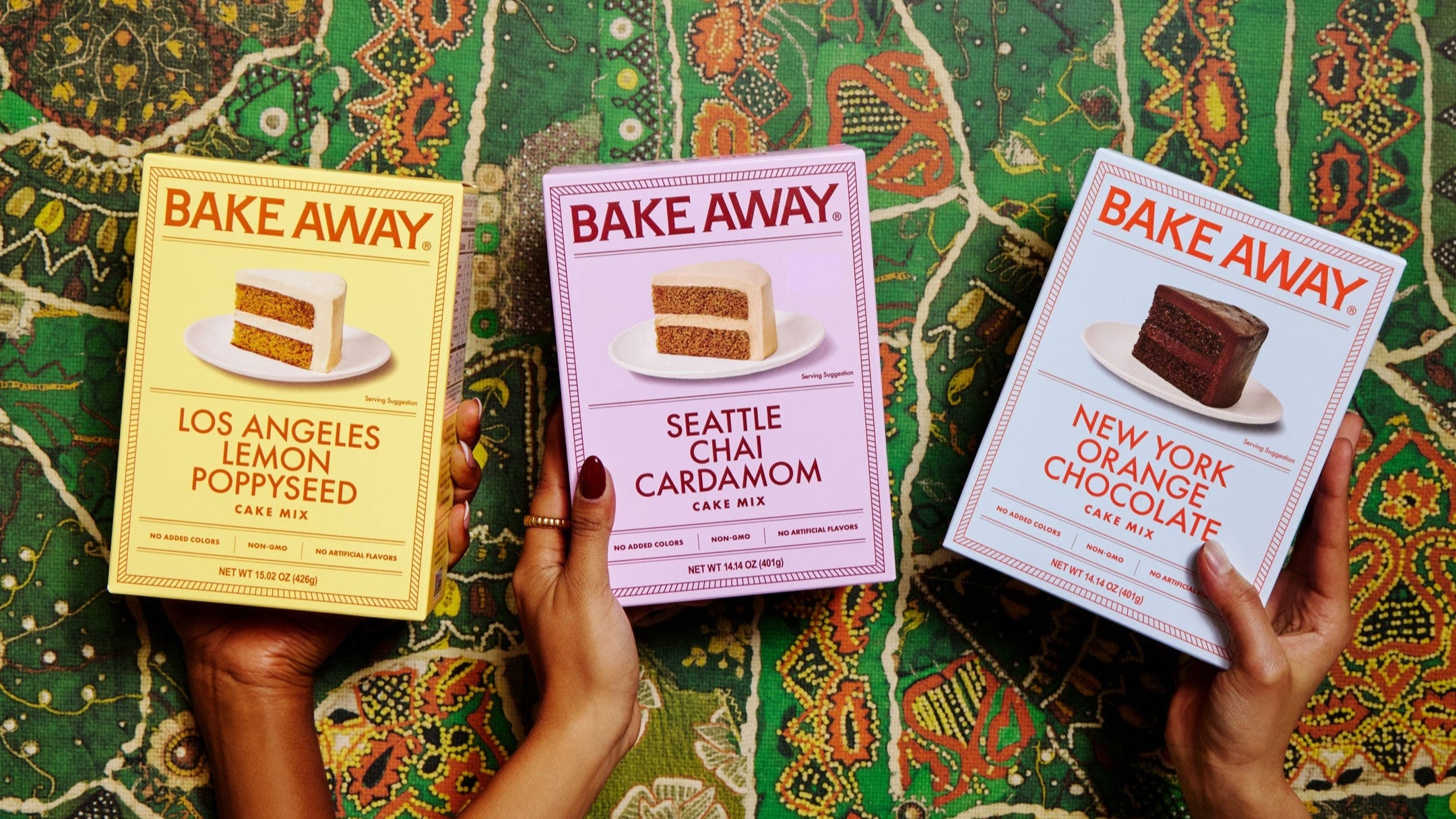 Three boxes of 'Bake Away' cake mixes held by hands on a patterned fabric background.